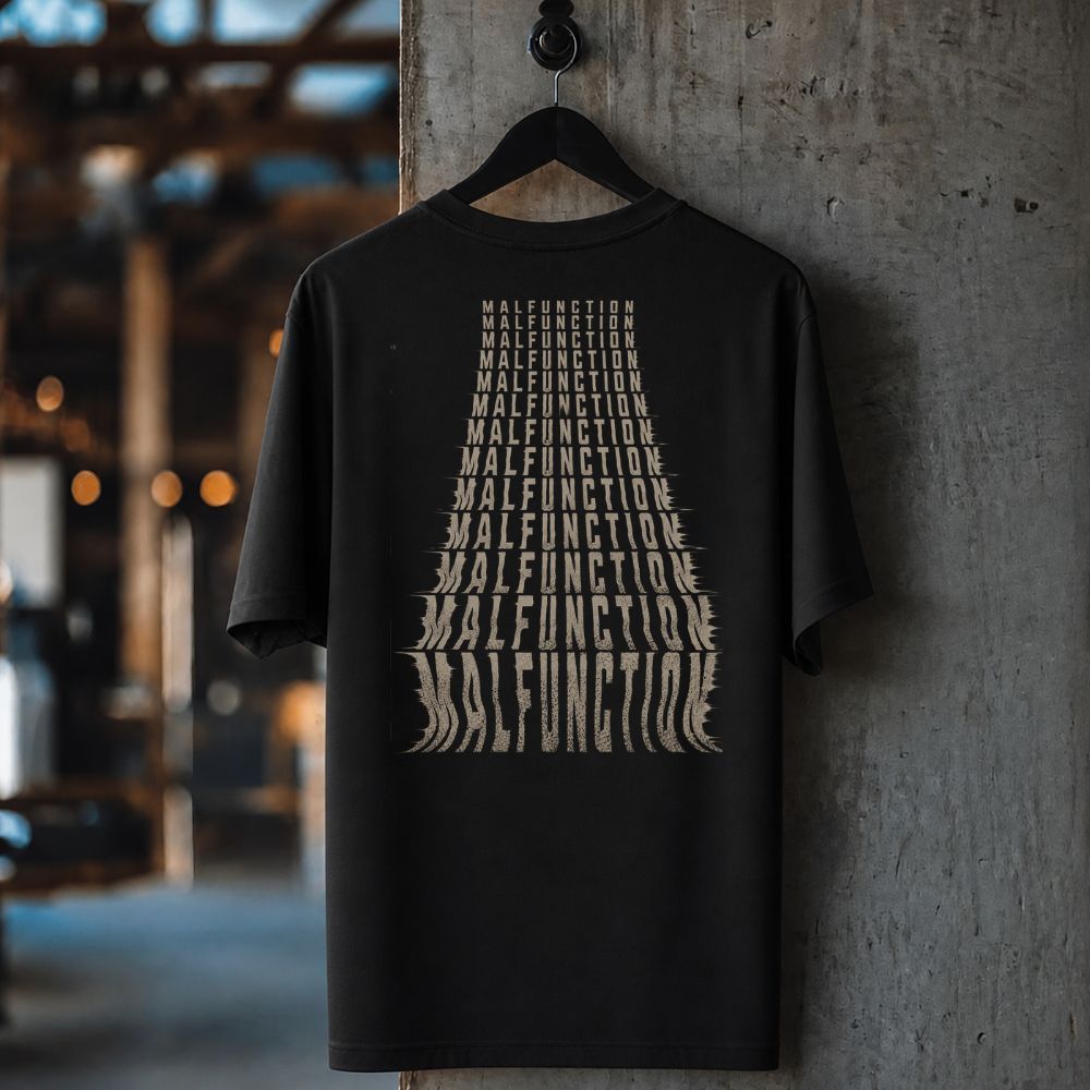 A black short-sleeve t-shirt hanging on a black hanger against a textured concrete pillar in an industrial setting. The shirt features a large graphic print on the front made of stacked, stylized beige text reading 'MALFUNCTION'.