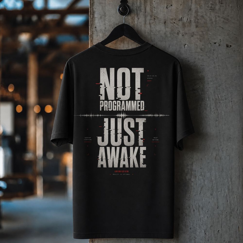 A black short-sleeve t-shirt hanging on a black hanger against a textured concrete pillar in an industrial loft setting. The shirt features a large white graphic text print reading 'NOT PROGRAMMED JUST AWAKE' with a glitch effect and small red accents.