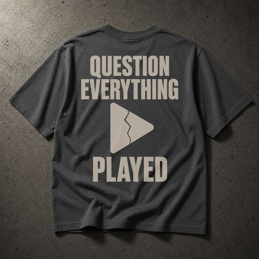 A dark gray t-shirt laid flat on a textured gray surface, showing the back with a light beige graphic that reads 'QUESTION EVERYTHING PLAYED' and a broken play triangle.