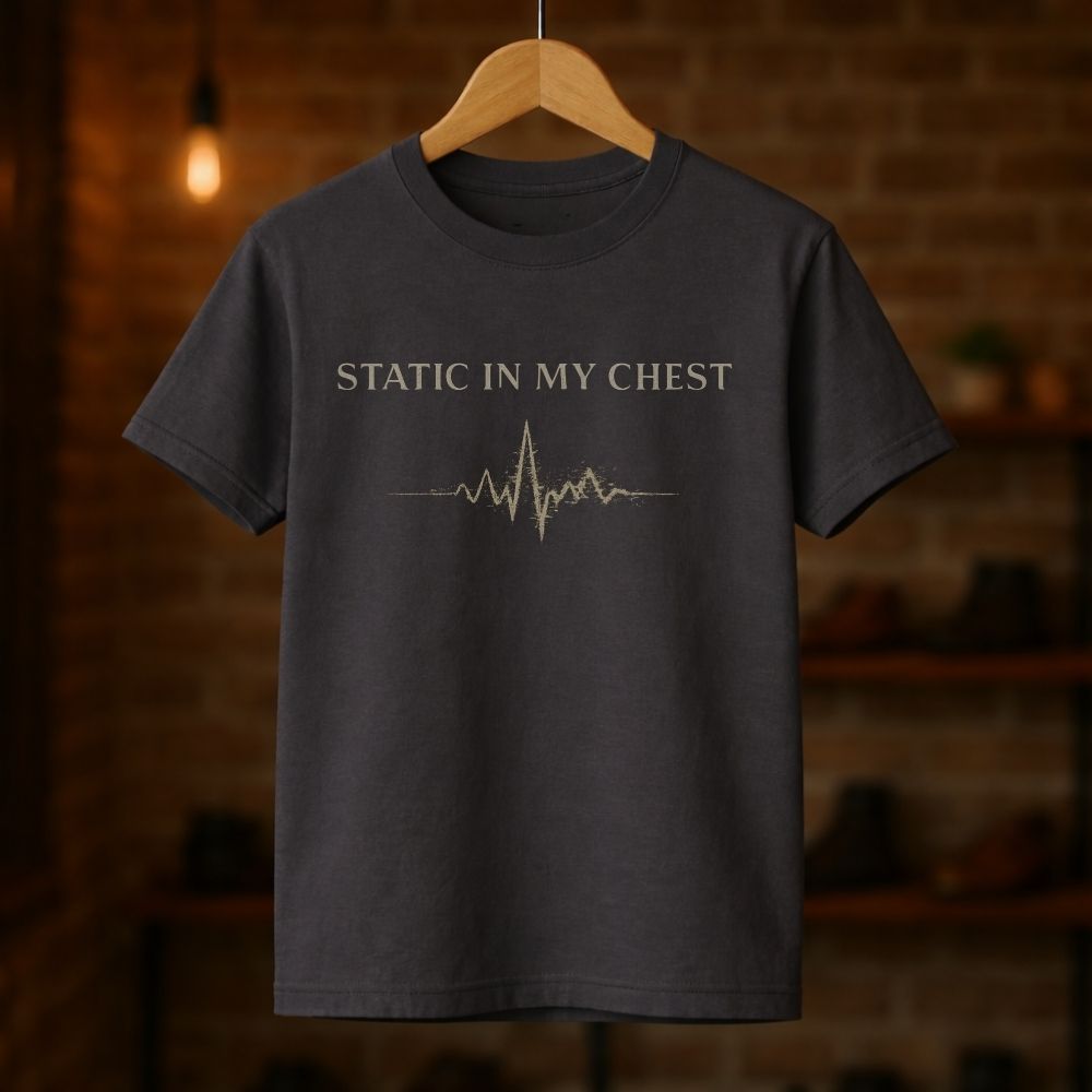 Dark gray t-shirt with 'Static in my chest' text and heartbeat line design on a wooden hanger against a blurred brick wall background.
