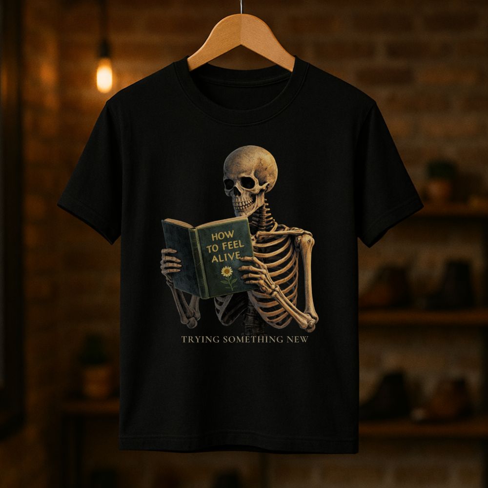 Black unisex graphic T-shirt hanging on a wooden hanger showing a skeleton reading "How to Feel Alive" with the text "Trying Something New"