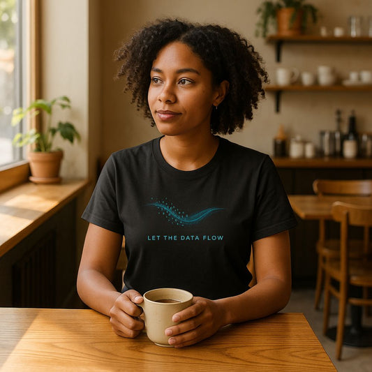 Woman wearing black “Let the Data Flow” T-shirt with blue binary wave design, sitting in cozy café holding coffee.