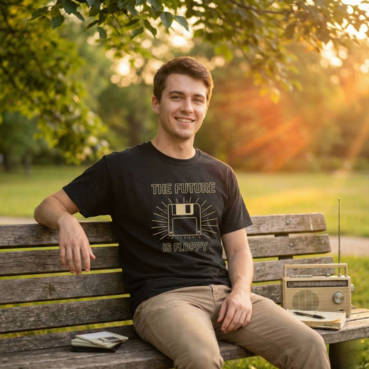 Male model wearing black “The Future Is Floppy” retro floppy disk t-shirt, sitting on a park bench with an analog radio and daylight sun rays in the background.