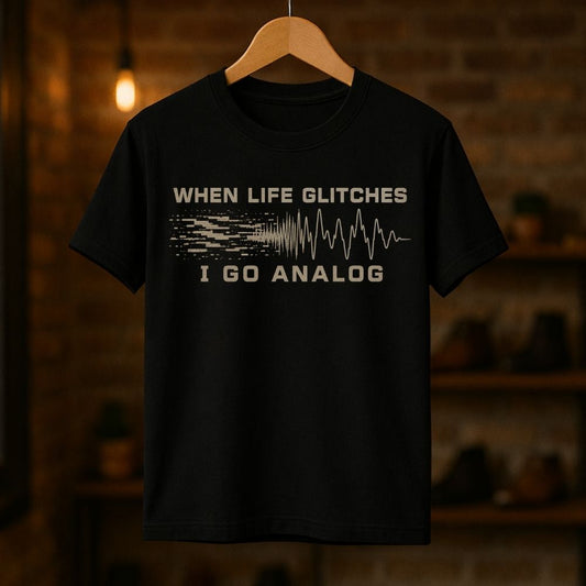 A black short-sleeve graphic t-shirt hanging on a wooden hanger against a warm, blurred brick wall background. The shirt features a distressed beige graphic that reads 'WHEN LIFE GLITCHES' above a pixelated signal wave transitioning into an analog waveform, with 'I GO ANALOG' printed below.
