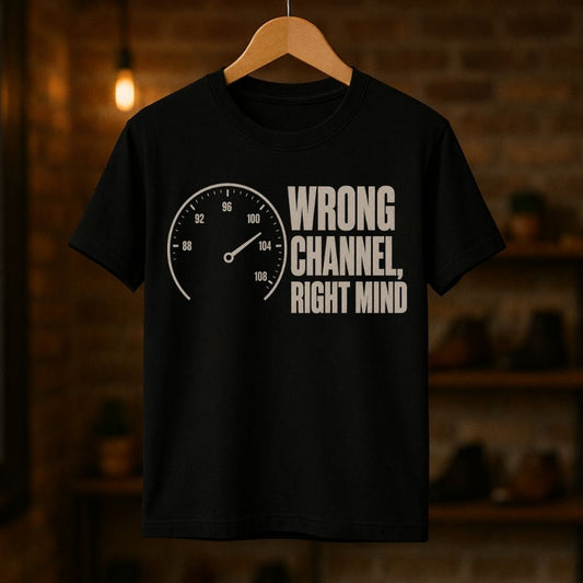 A black short-sleeve t-shirt hanging on a wooden hanger against a warm, blurred brick wall background. The shirt features a white graphic print of a speedometer on the left and the text 'WRONG CHANNEL, RIGHT MIND' on the right.