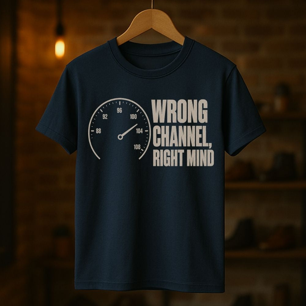 A navy blue graphic t-shirt hanging on a wooden hanger against a warm, blurred brick wall background. The shirt features a white graphic of a speedometer next to the text 'WRONG CHANNEL, RIGHT MIND'.