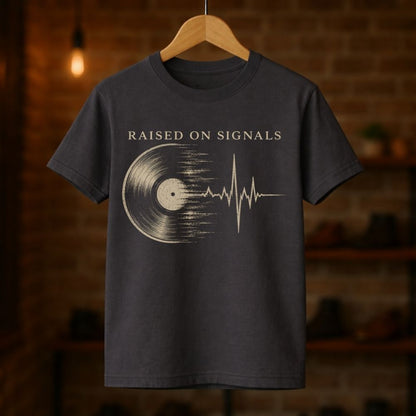 Dark gray t-shirt with 'Raised on Signals' design on a wooden hanger against a blurred brick wall background.