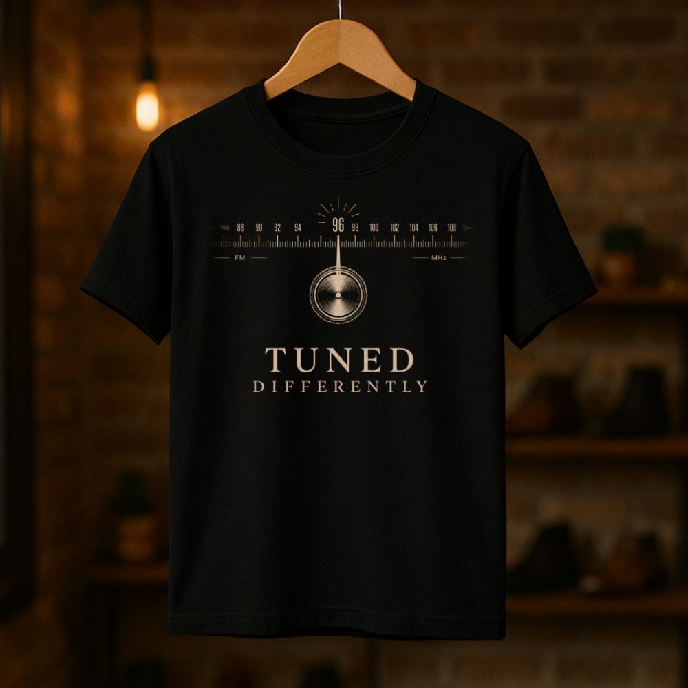 Black t-shirt with radio dial graphic and 'Tuned Differently' text on a wooden hanger against a blurred brick wall background.