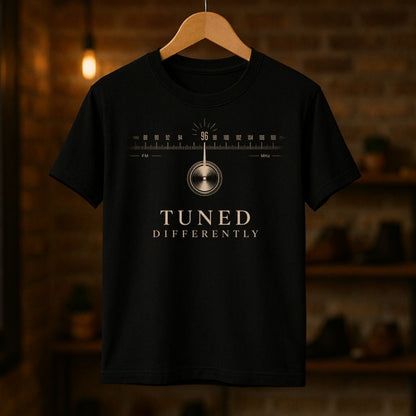 Black t-shirt with radio dial graphic and 'Tuned Differently' text on a wooden hanger against a blurred brick wall background.