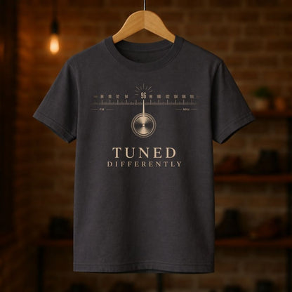 Black t-shirt with 'Tuned Differently' design on a wooden hanger against a blurred brick wall background.