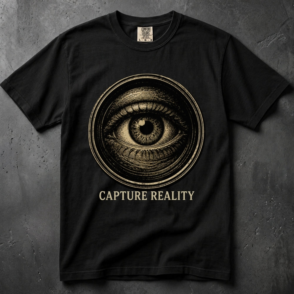 A black short-sleeve crewneck t-shirt featuring a large circular graphic of an all-seeing eye in a sepia tone, with the text 'CAPTURE REALITY' printed below in a matching serif font. The shirt is displayed flat against a dark, textured gray background.