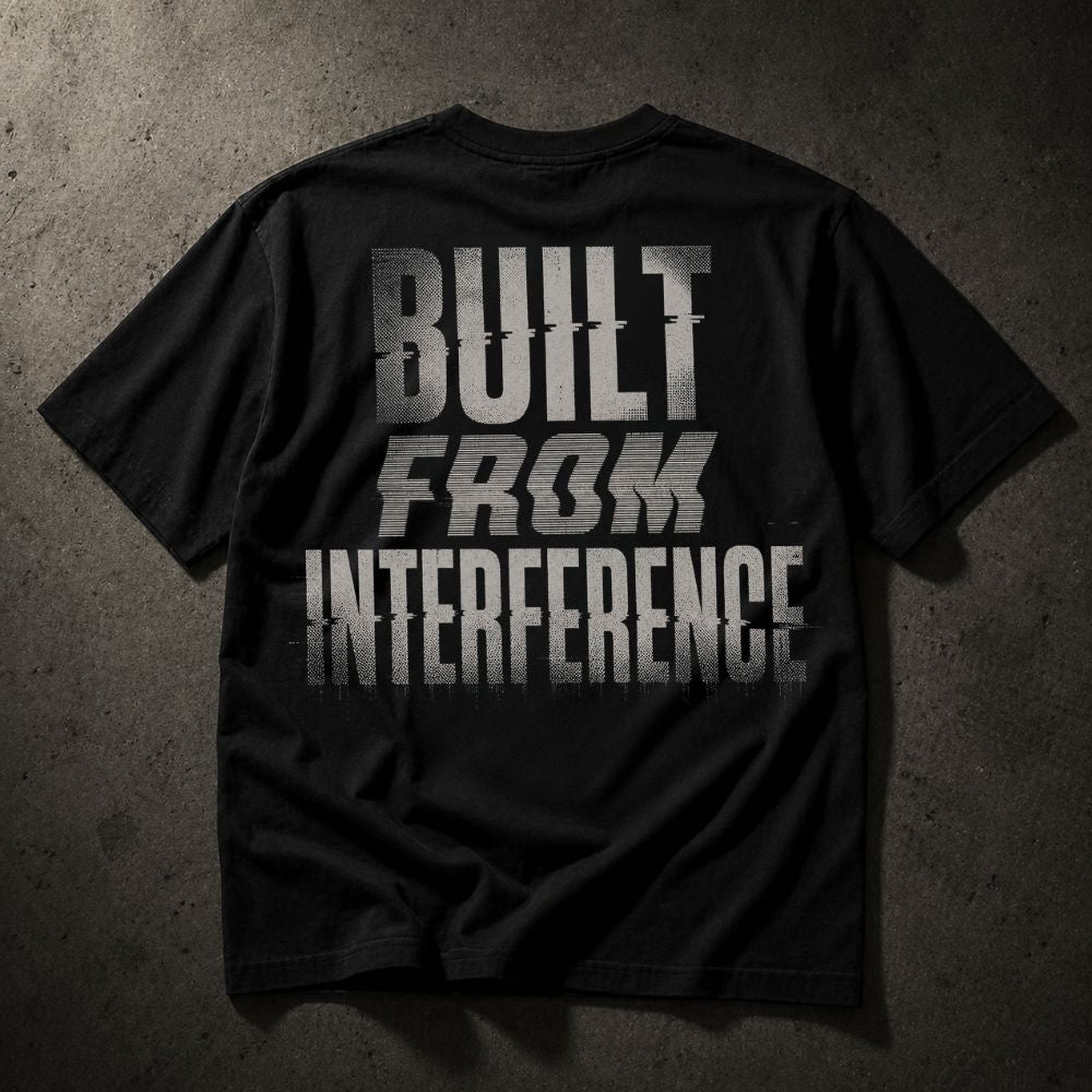 A black short-sleeve t-shirt laid flat on a textured dark gray background, featuring the phrase 'BUILT FROM INTERFERENCE' printed in large distressed white letters across the back.