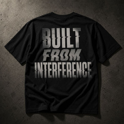 A black short-sleeve t-shirt laid flat on a textured dark gray background, featuring the phrase 'BUILT FROM INTERFERENCE' printed in large distressed white letters across the back.