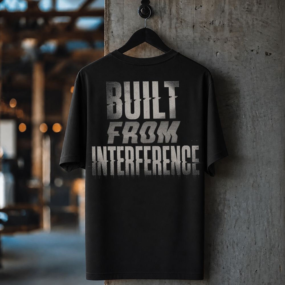 A black short-sleeve t-shirt hanging on a black hanger against a textured concrete pillar, featuring a distressed silver graphic print that reads 'BUILT FROM INTERFERENCE'.