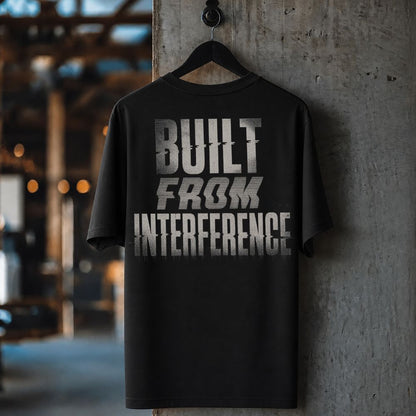 A black short-sleeve t-shirt hanging on a black hanger against a textured concrete pillar, featuring a distressed silver graphic print that reads 'BUILT FROM INTERFERENCE'.