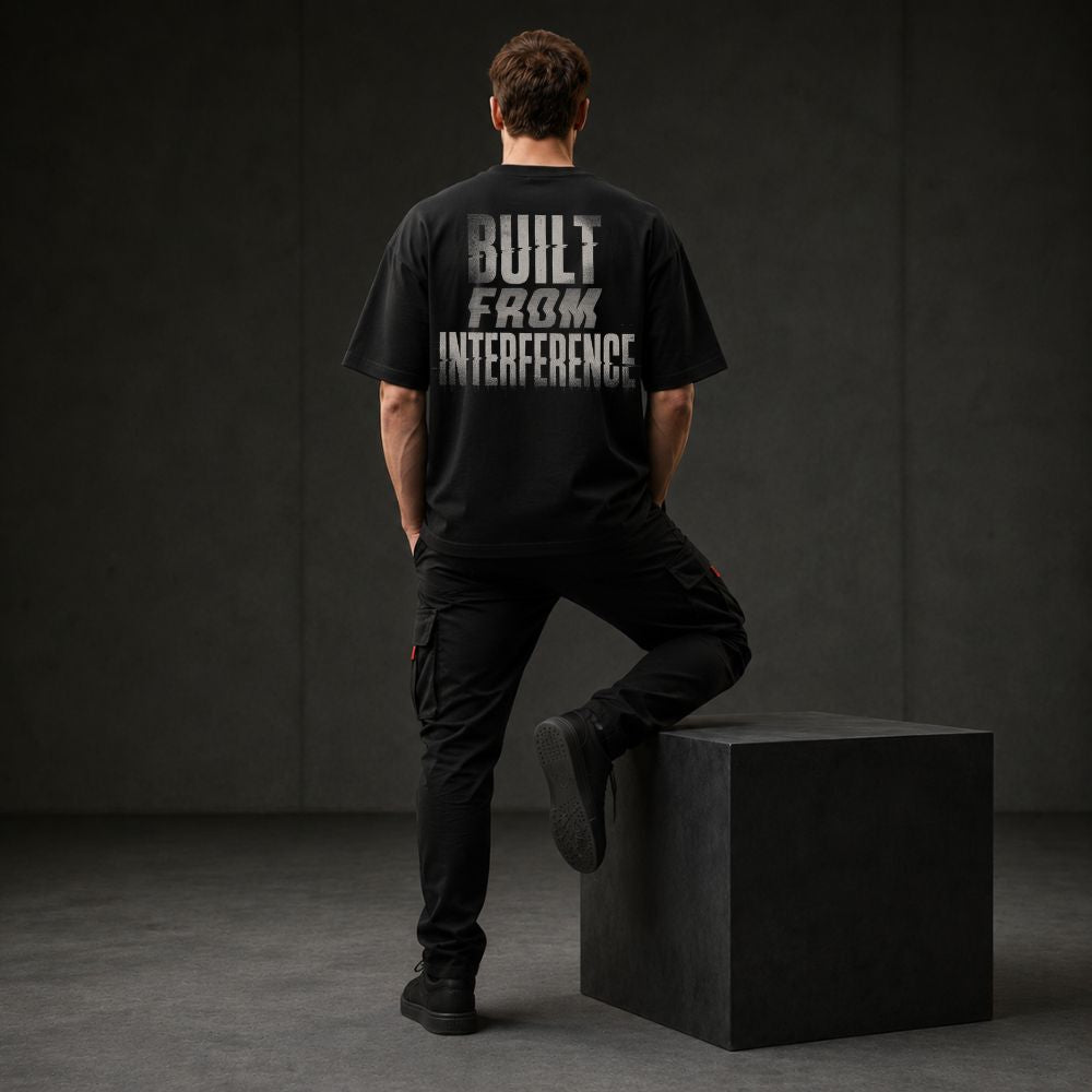 A man stands with his back to the camera wearing a black oversized T-shirt with a distressed white graphic reading 'BUILT FROM INTERFERENCE' on the back, paired with black cargo pants and black shoes, posed on a dark gray cube in a studio setting.