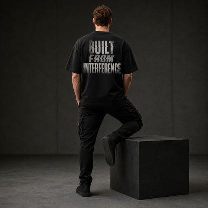 A man stands with his back to the camera wearing a black oversized T-shirt with a distressed white graphic reading 'BUILT FROM INTERFERENCE' on the back, paired with black cargo pants and black shoes, posed on a dark gray cube in a studio setting.