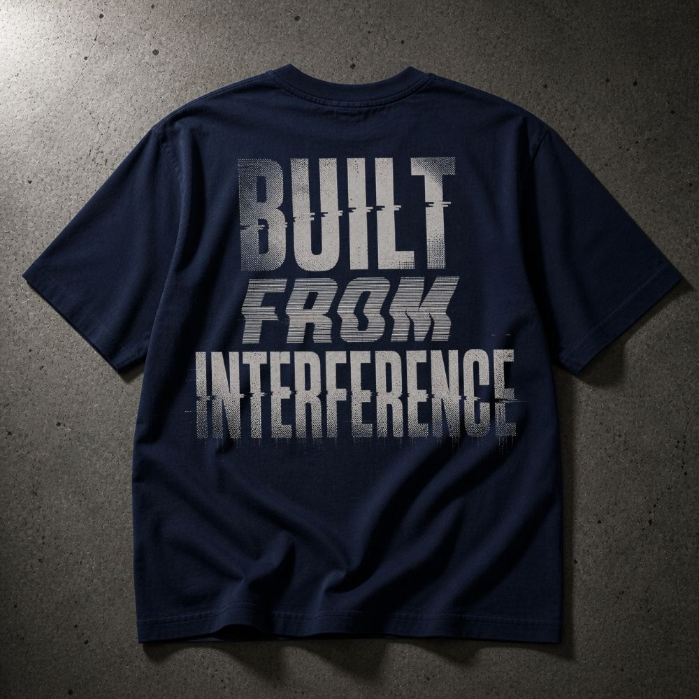 A navy blue short-sleeve t-shirt laid flat on a textured gray concrete surface, featuring a distressed white graphic print on the back that reads 'BUILT FROM INTERFERENCE'.