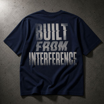 A navy blue short-sleeve t-shirt laid flat on a textured gray concrete surface, featuring a distressed white graphic print on the back that reads 'BUILT FROM INTERFERENCE'.