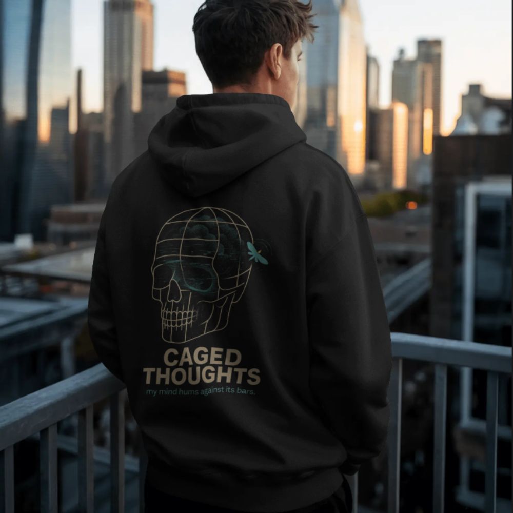 Model wearing black Caged Thoughts hoodie with skull graphic on the back, standing on a city rooftop at dusk