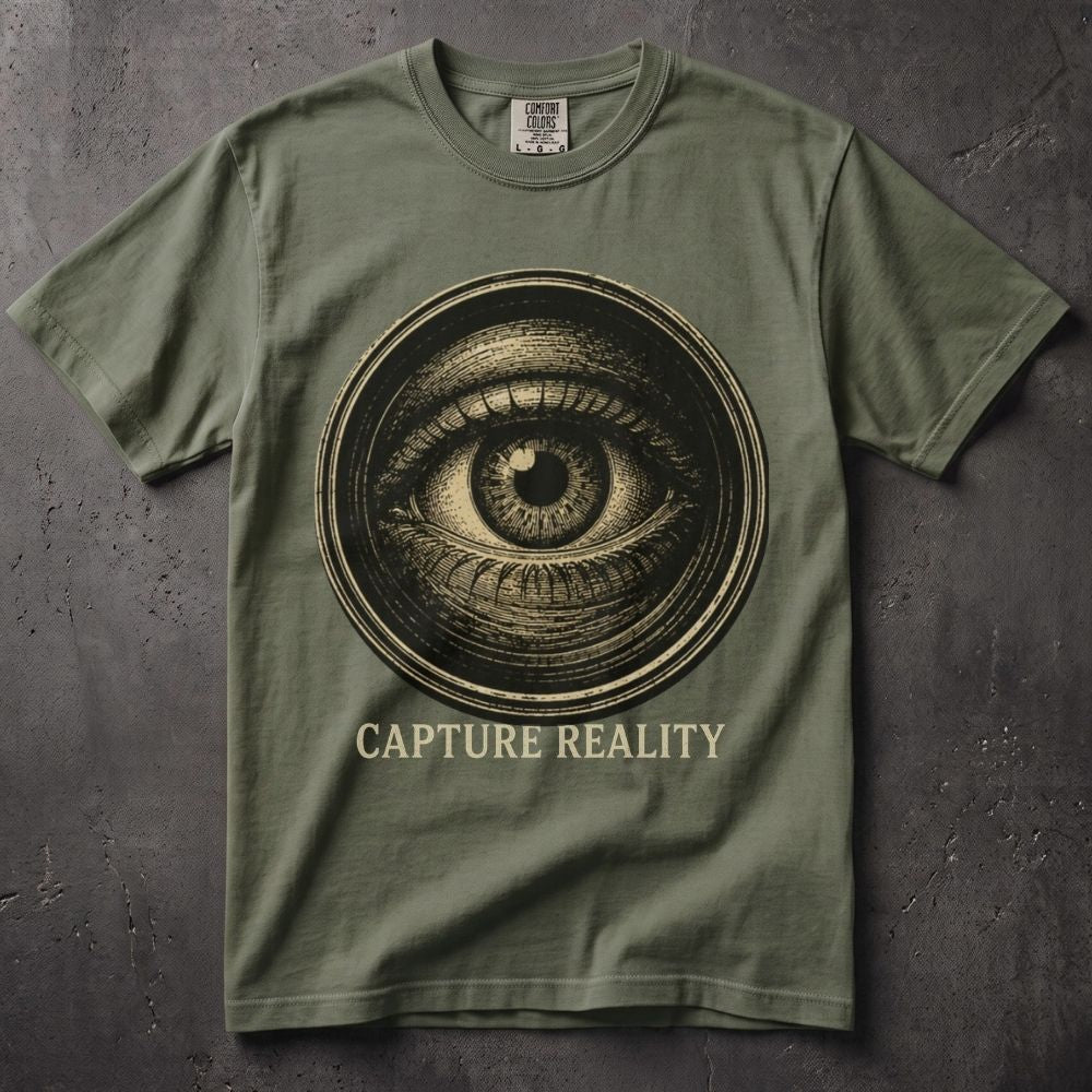 An olive green crewneck t-shirt featuring a large circular graphic of an all-seeing eye in a vintage sepia tone, with the text 'CAPTURE REALITY' printed below in a matching beige font. The shirt is laid flat on a dark, textured concrete-like surface.