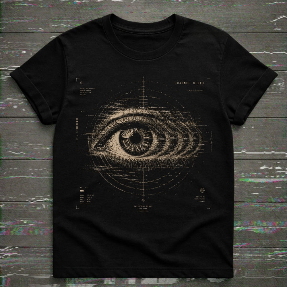 A black short-sleeve crewneck t-shirt featuring a detailed beige graphic of a human eye with technical blueprint markings and the text 'CHANNEL BLEED' on the chest, displayed flat on a dark wood plank background.