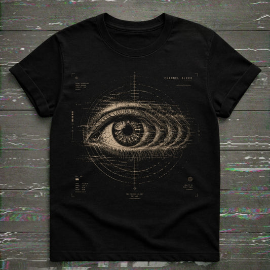 A black short-sleeve crewneck t-shirt featuring a detailed beige graphic of a human eye with technical blueprint markings and the text 'CHANNEL BLEED' on the chest, displayed flat on a dark wood plank background.