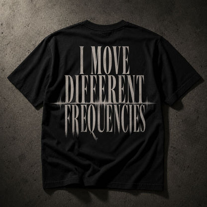 A black short-sleeve t-shirt laid flat on a dark textured concrete background, featuring a distressed white graphic print on the back that reads 'I MOVE DIFFERENT FREQUENCIES' with a soundwave line detail.