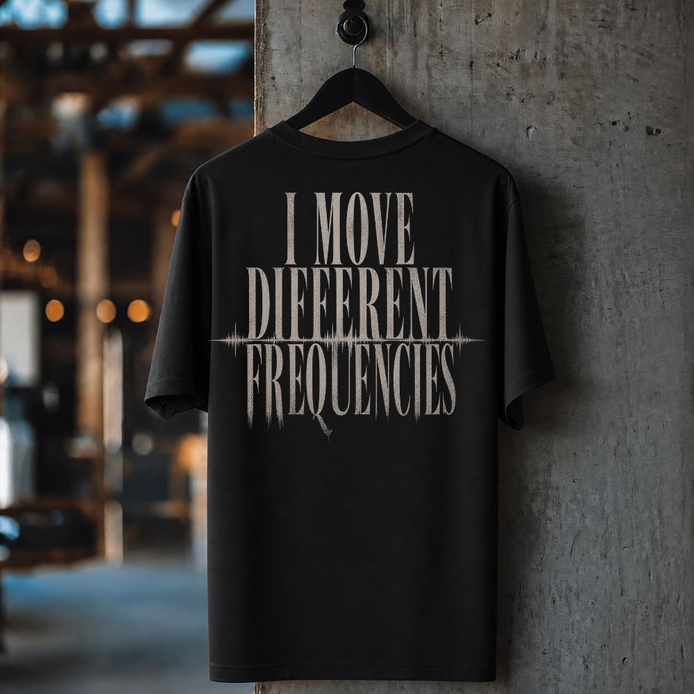 A black short-sleeve t-shirt hanging on a black hanger against a textured concrete pillar, featuring the phrase 'I MOVE DIFFERENT FREQUENCIES' printed in a distressed white font on the back.