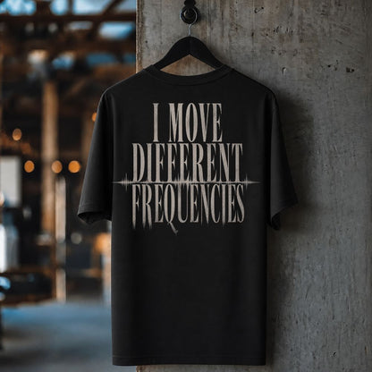 A black short-sleeve t-shirt hanging on a black hanger against a textured concrete pillar, featuring the phrase 'I MOVE DIFFERENT FREQUENCIES' printed in a distressed white font on the back.