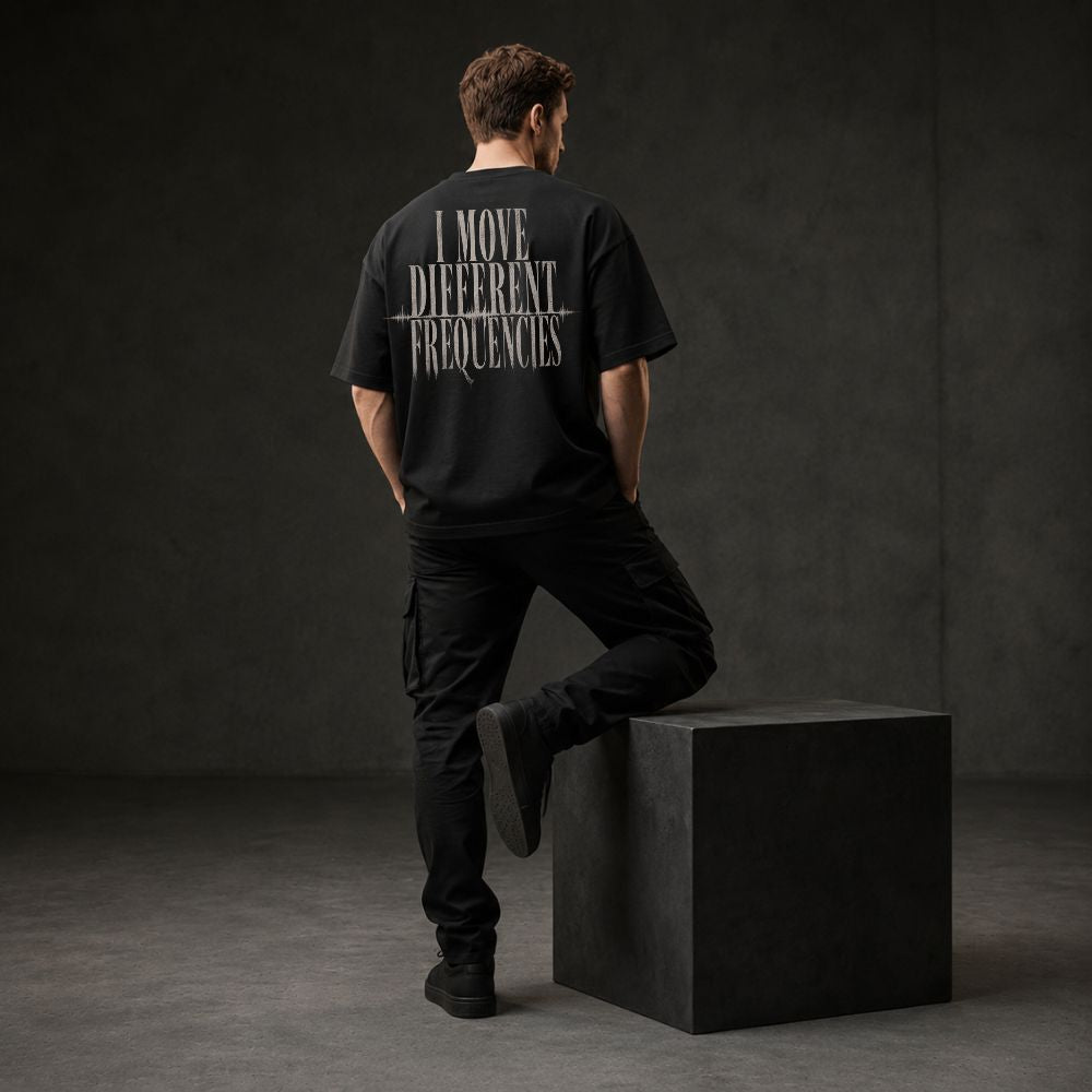 A man stands with his back to the camera wearing a black t-shirt with white text reading 'I MOVE DIFFERENT FREQUENCIES' across the back, paired with black cargo pants and standing on a dark gray block in a studio setting.