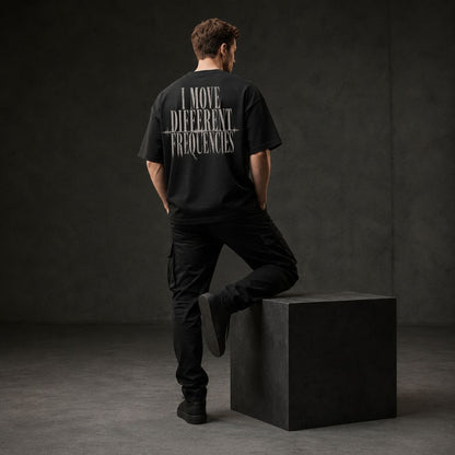 A man stands with his back to the camera wearing a black t-shirt with white text reading 'I MOVE DIFFERENT FREQUENCIES' across the back, paired with black cargo pants and standing on a dark gray block in a studio setting.