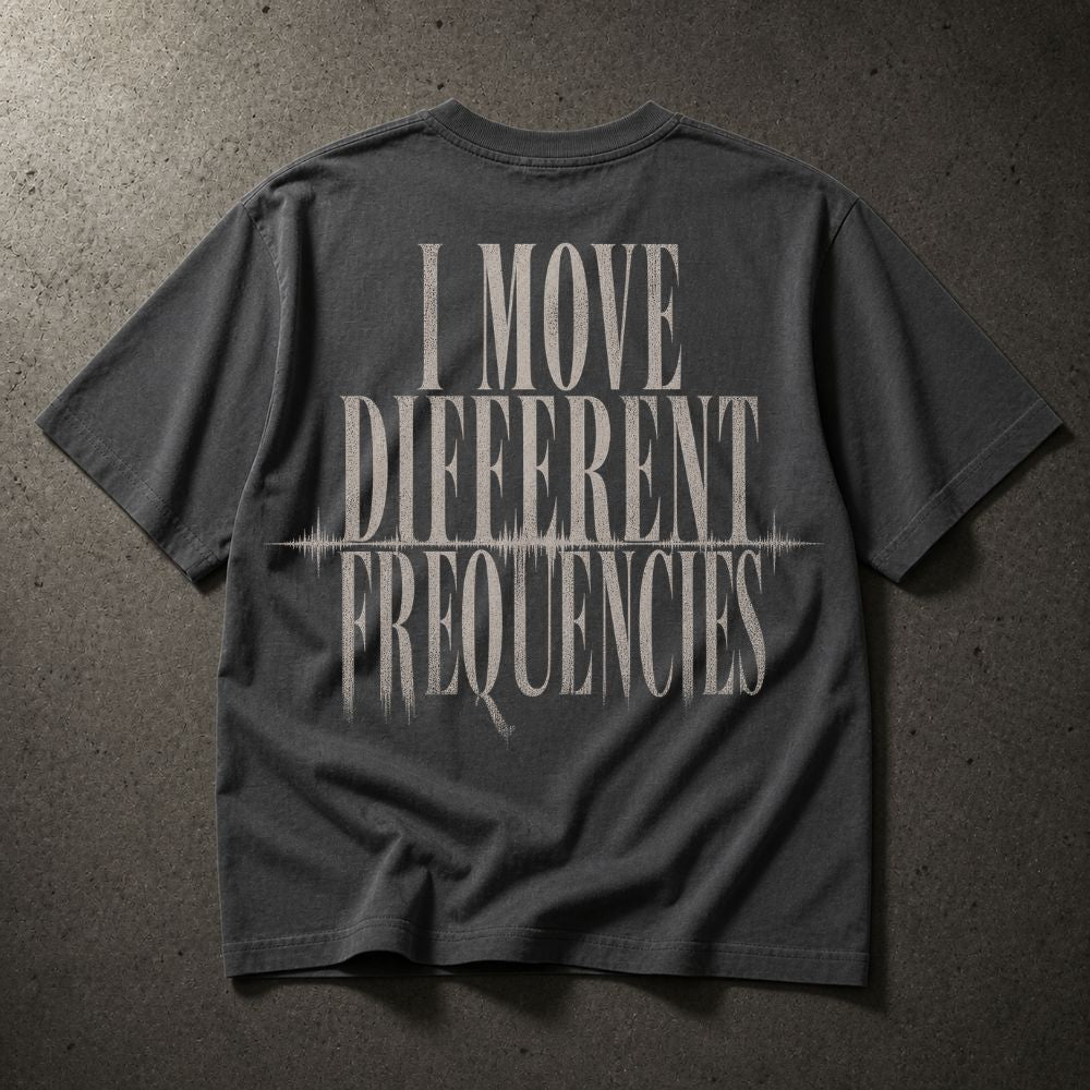 A dark gray t-shirt laid flat on a textured gray concrete surface, featuring a distressed white graphic print on the back that reads 'I MOVE DIFFERENT FREQUENCIES' with a soundwave line detail.