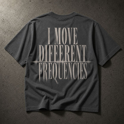 A dark gray t-shirt laid flat on a textured gray concrete surface, featuring a distressed white graphic print on the back that reads 'I MOVE DIFFERENT FREQUENCIES' with a soundwave line detail.
