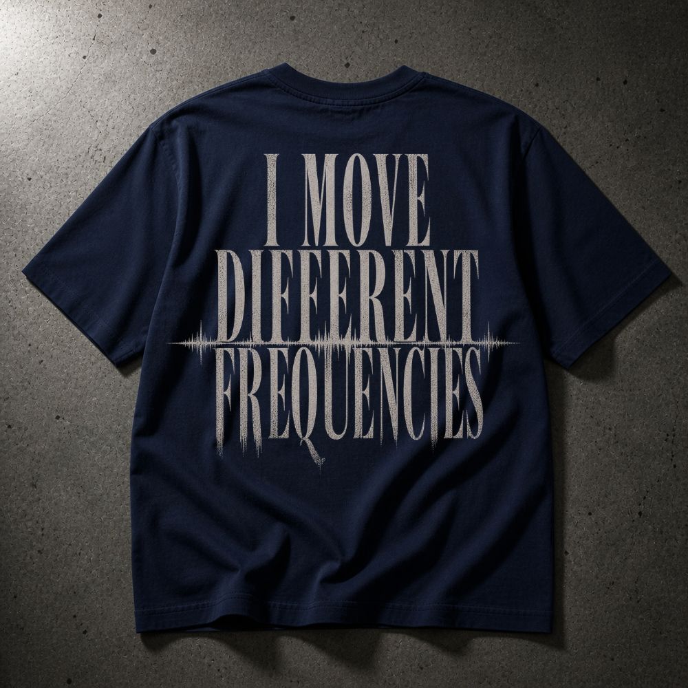 A navy blue short-sleeve t-shirt laid flat on a textured gray concrete surface, featuring a large distressed white graphic on the back that reads 'I MOVE DIFFERENT FREQUENCIES' with a soundwave line detail.