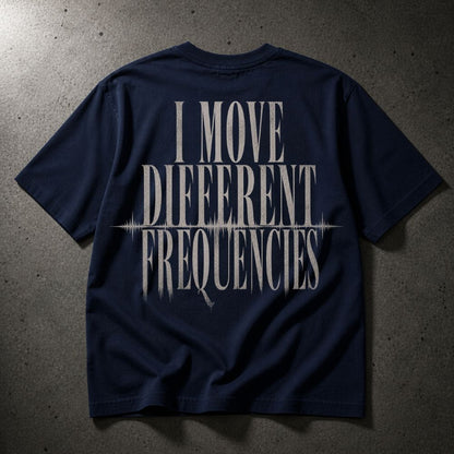 A navy blue short-sleeve t-shirt laid flat on a textured gray concrete surface, featuring a large distressed white graphic on the back that reads 'I MOVE DIFFERENT FREQUENCIES' with a soundwave line detail.