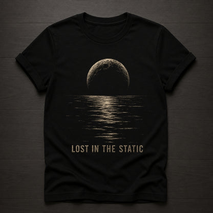 A black short-sleeve t-shirt laid flat on a dark wood background, featuring a distressed beige graphic of a moon over water with the text 'LOST IN THE STATIC' printed below.