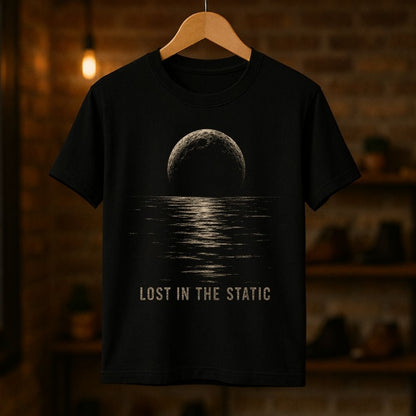 A black short-sleeve t-shirt hanging on a wooden hanger against a warm, blurred brick wall background. The shirt features a graphic print of a dark moon over water with the text 'LOST IN THE STATIC' printed below in a distressed beige font.