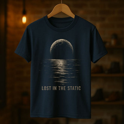 A navy blue short-sleeve t-shirt hanging on a wooden hanger against a warm, blurred brick wall background. The shirt features a graphic print of a moon over water with the text 'LOST IN THE STATIC' printed below in a distressed beige font.