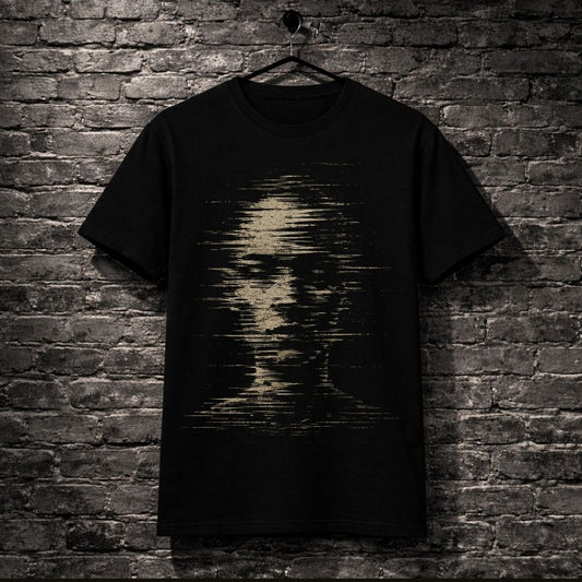 A black short-sleeve t-shirt hanging on a black hanger against a dark gray brick wall. The shirt features a distressed, pixelated graphic of a human face in a muted beige or tan color on the front.