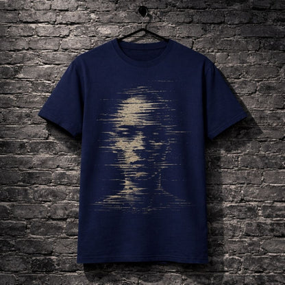 A navy blue short-sleeve t-shirt hanging on a black hanger against a dark gray brick wall. The front of the shirt features a distressed, pixelated graphic of a face in a light beige or cream color.