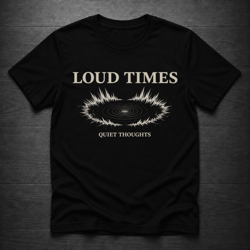 A black short-sleeve t-shirt laid flat on a dark wood background, featuring a cream-colored graphic with the text 'LOUD TIMES' and 'QUIET THOUGHTS' surrounding a soundwave design.