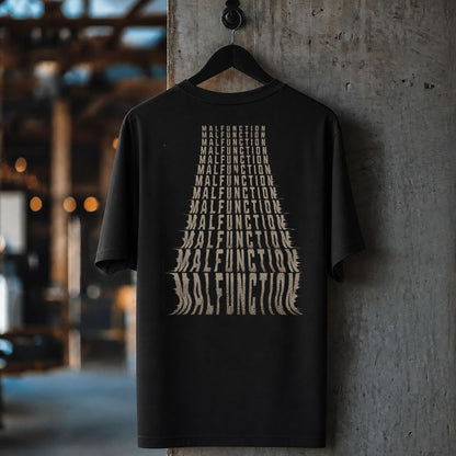 A black short-sleeve t-shirt hanging on a black hanger against a textured concrete pillar in an industrial setting. The shirt features a large graphic print on the front made of stacked, stylized beige text reading 'MALFUNCTION'.