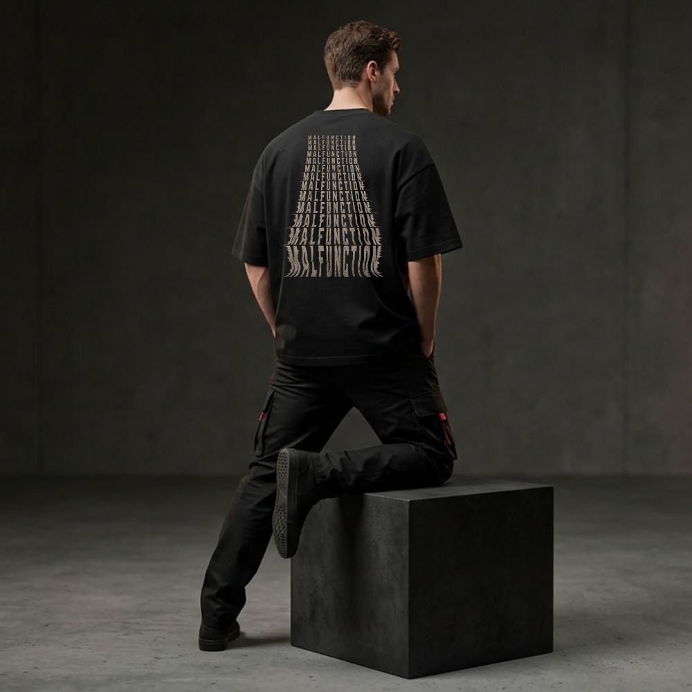 A man wearing a black oversized T-shirt with a large white graphic print on the back, posed from behind sitting on a black block in a dark studio setting.