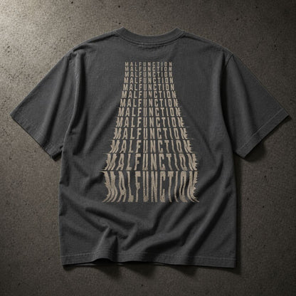 A dark gray t-shirt laid flat on a textured gray background, showing the back with a large beige graphic of the word 'MALFUNCTION' repeated in a converging triangle pattern.