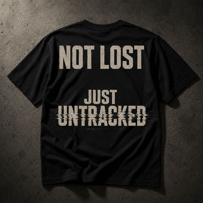 A black short-sleeve t-shirt laid flat on a dark textured background, featuring the distressed beige text 'NOT LOST JUST UNTRACKED' printed on the back.