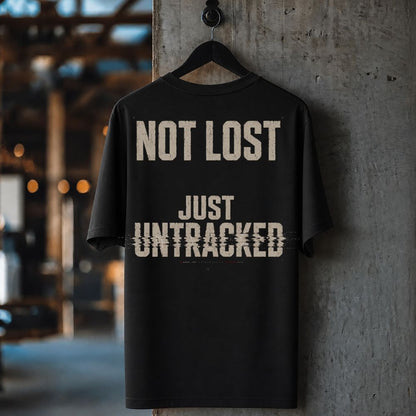 A black short-sleeve t-shirt hanging on a black hanger against a textured concrete pillar, featuring the distressed beige text 'NOT LOST JUST UNTRACKED' on the back.