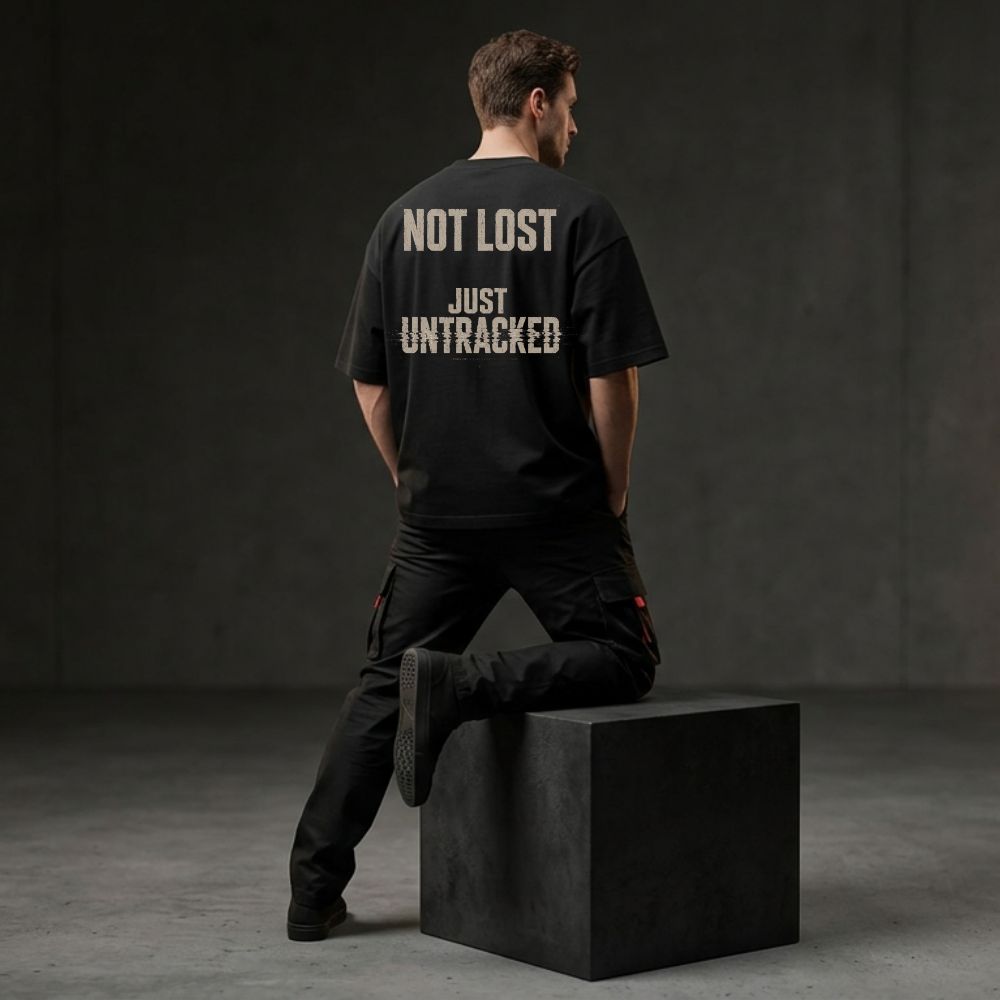 A man wearing a black t-shirt with the text 'NOT LOST JUST UNTRACKED' printed in white on the back, posing from behind while sitting on a black block in a dark studio setting.
