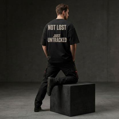 A man wearing a black t-shirt with the text 'NOT LOST JUST UNTRACKED' printed in white on the back, posing from behind while sitting on a black block in a dark studio setting.