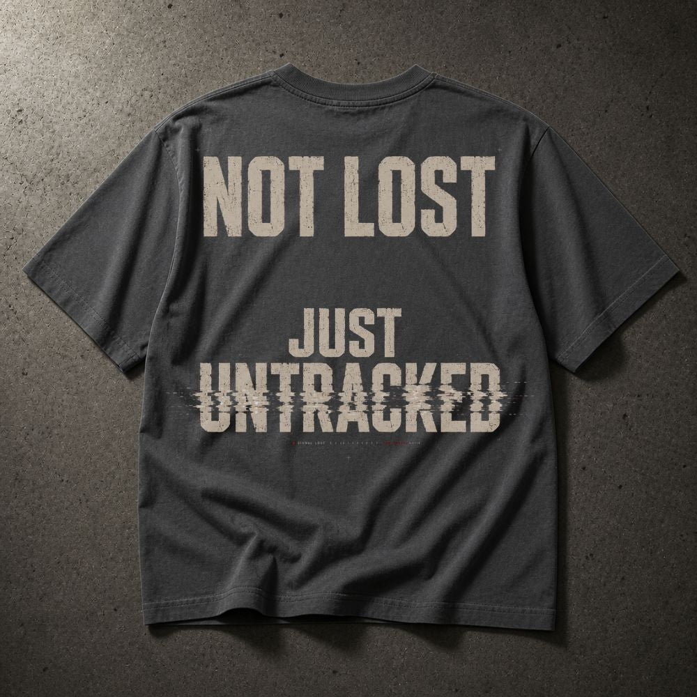 A dark charcoal gray t-shirt laid flat on a textured gray concrete surface, featuring the distressed beige text 'NOT LOST JUST UNTRACKED' printed on the back.